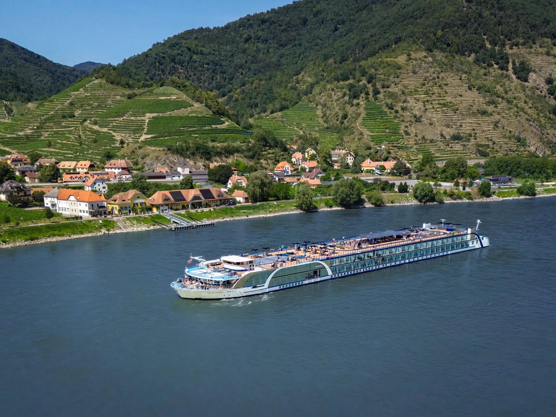 Long river cruise ship passing a village along a green mountainside riverbank