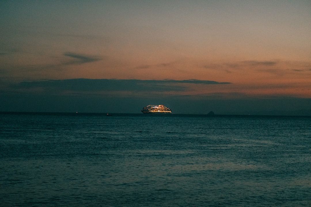 A brightly lit cruise ship sits on the dark ocean horizon under a dusky, golden-hued sunset sky.