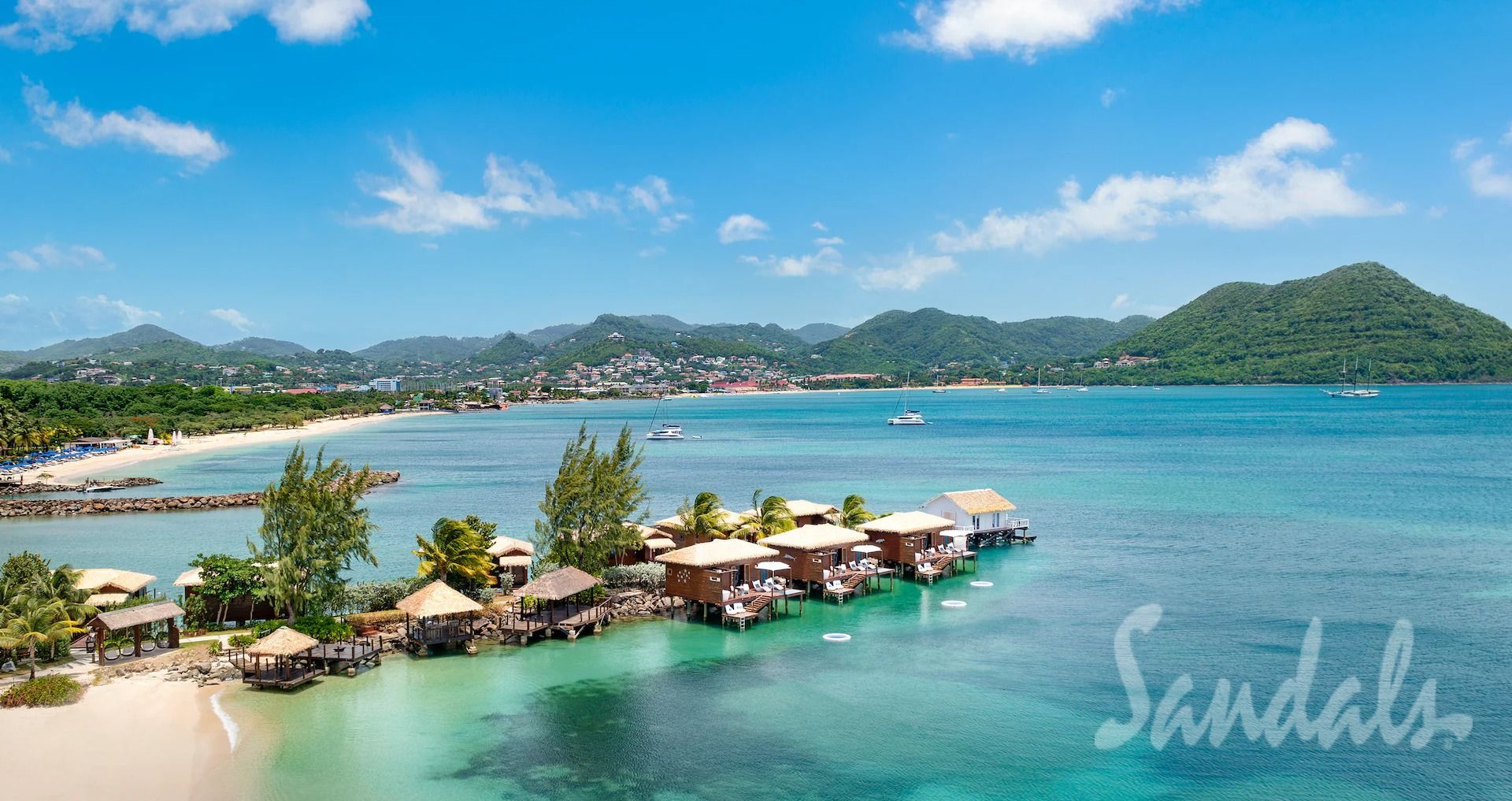 Tropical resort over turquoise bay with overwater huts, palm trees, and green hills under a clear blue sky