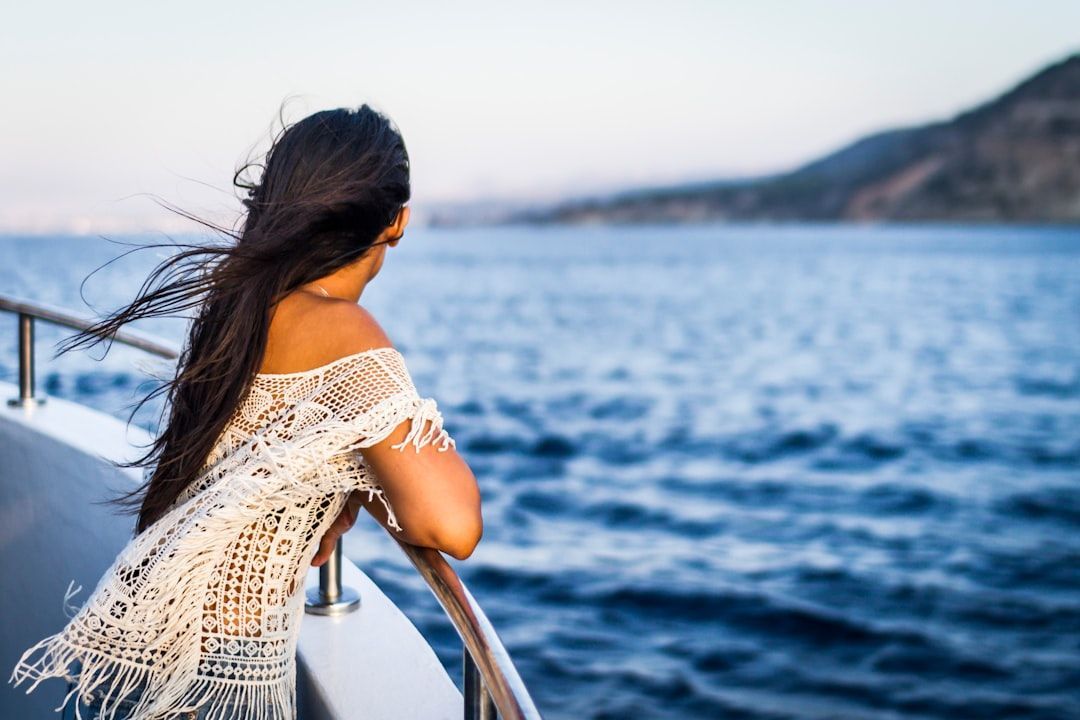 A person with long dark hair, wearing a white crochet top, leans over a boat railing, looking out toward distant mountains.