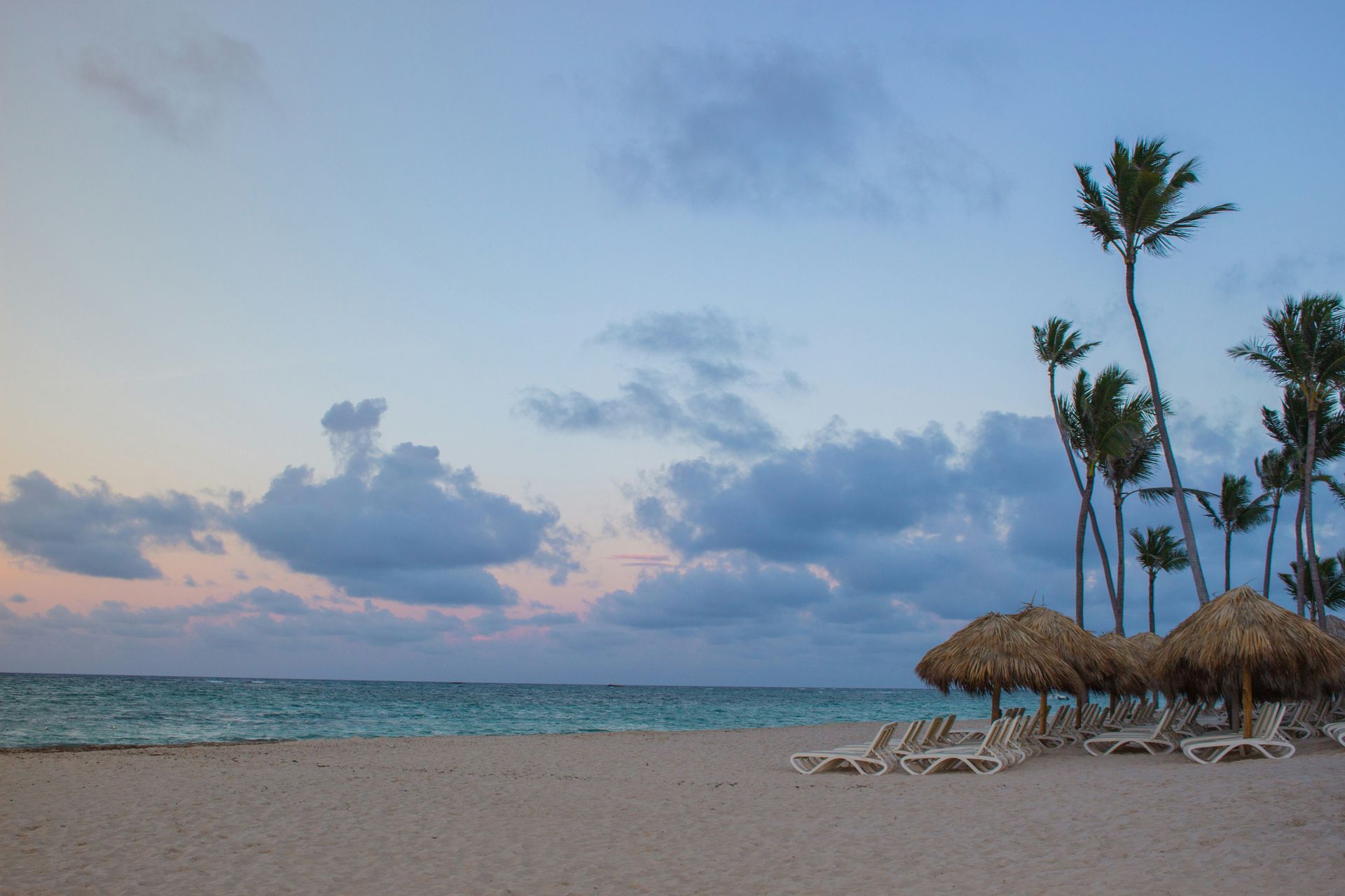 Quiet tropical beach at sunset with lounge chairs, palm trees, and a calm ocean horizon.