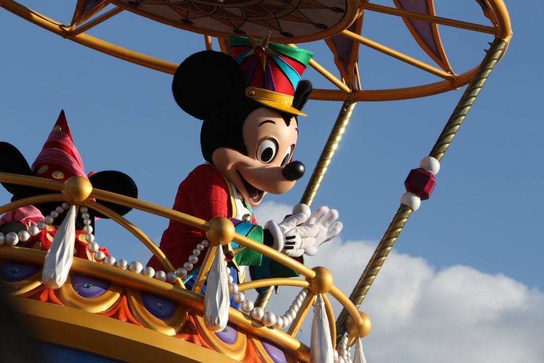 Mickey Mouse in a colorful parade outfit waves from a decorated float against a blue sky.
