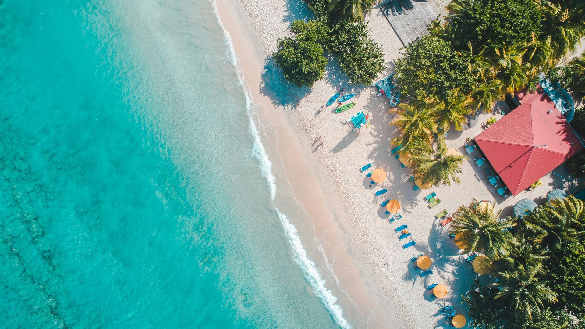 Aerial view of a tropical beach with turquoise water, white sand, palm trees, and red-roofed huts