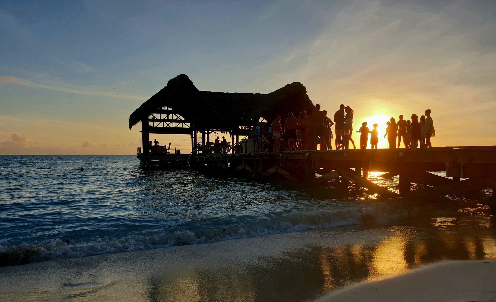 Silhouetted people on a pier at sunset over the ocean, with a thatched-roof hut and glowing sky