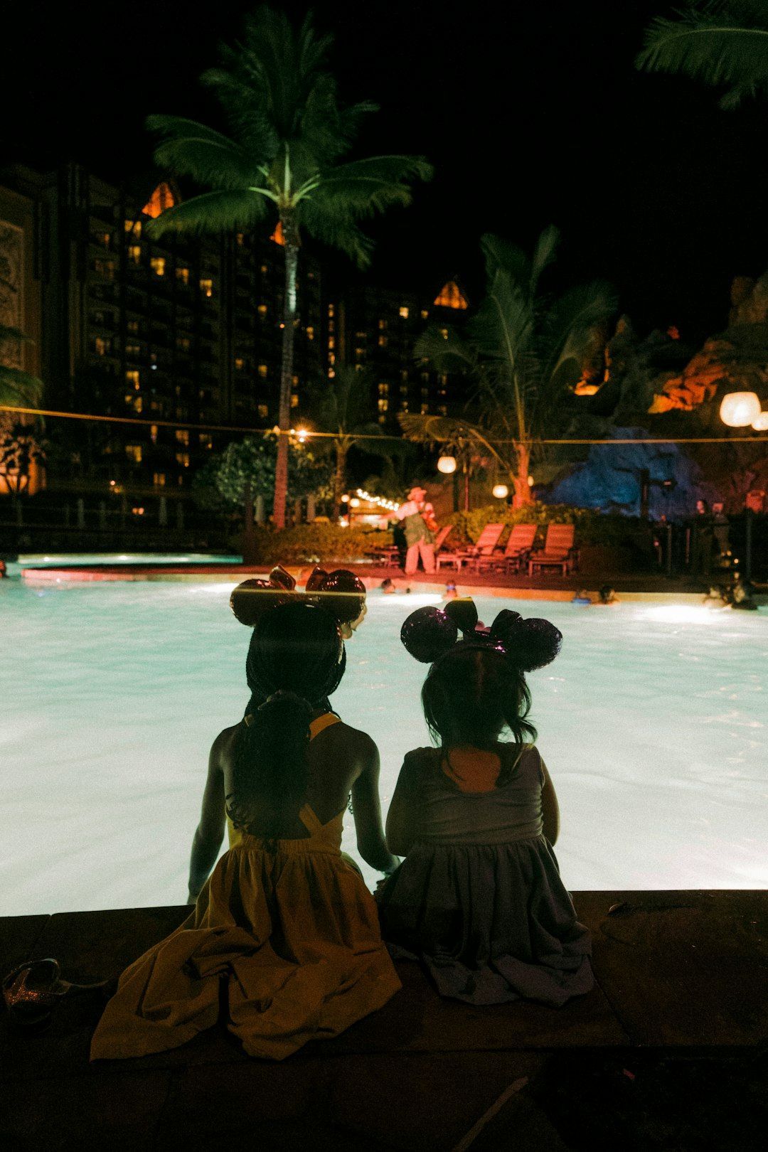 Two children sit by a glowing pool at night, with palm trees and lit resort buildings in the background.