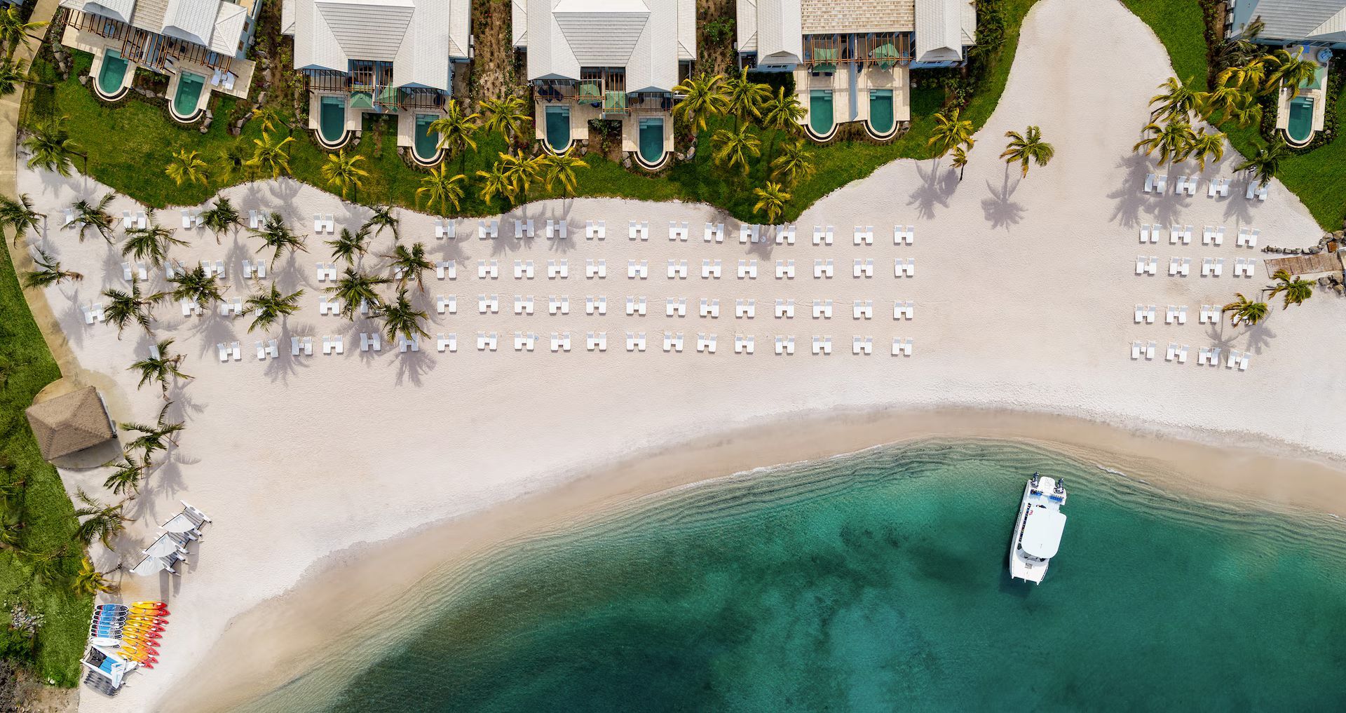 Aerial view of a tropical beach with lounge chairs, palm trees, resort villas, and a small boat in turquoise water