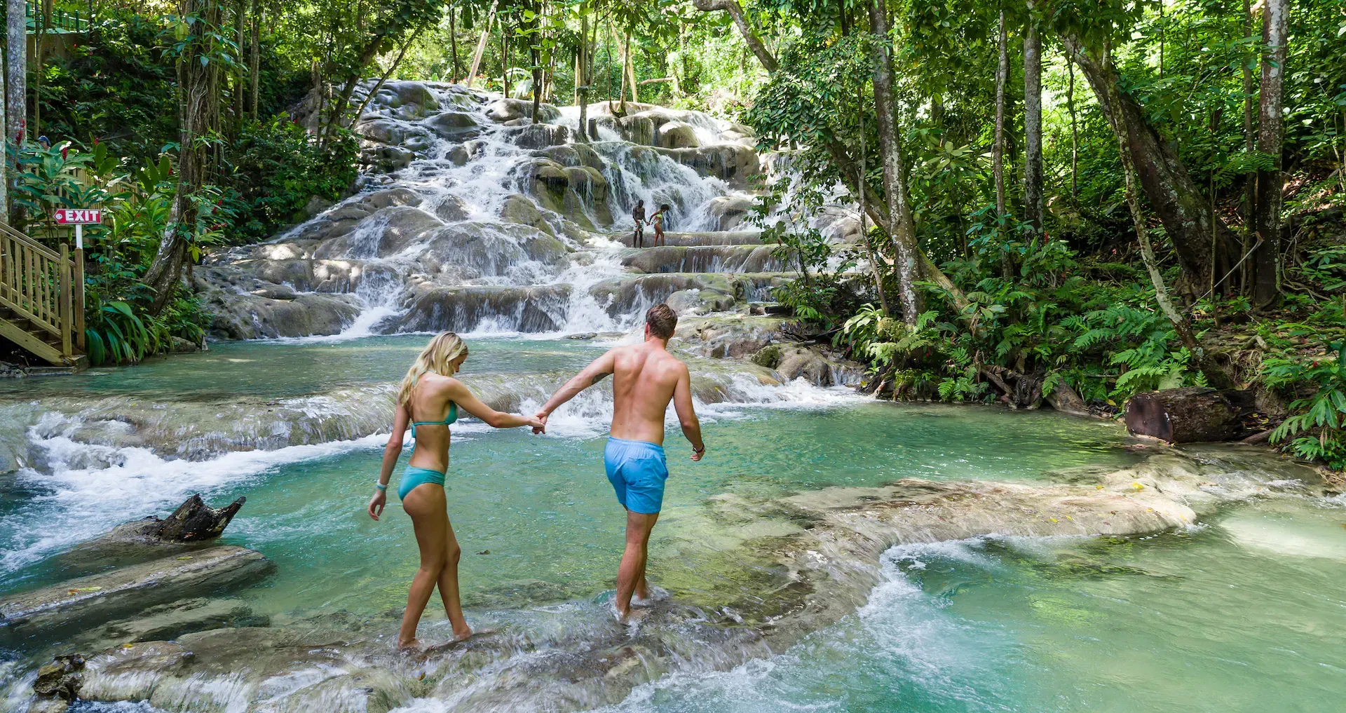 Two people walking through shallow turquoise water toward a tiered tropical waterfall in a lush, green forest.
