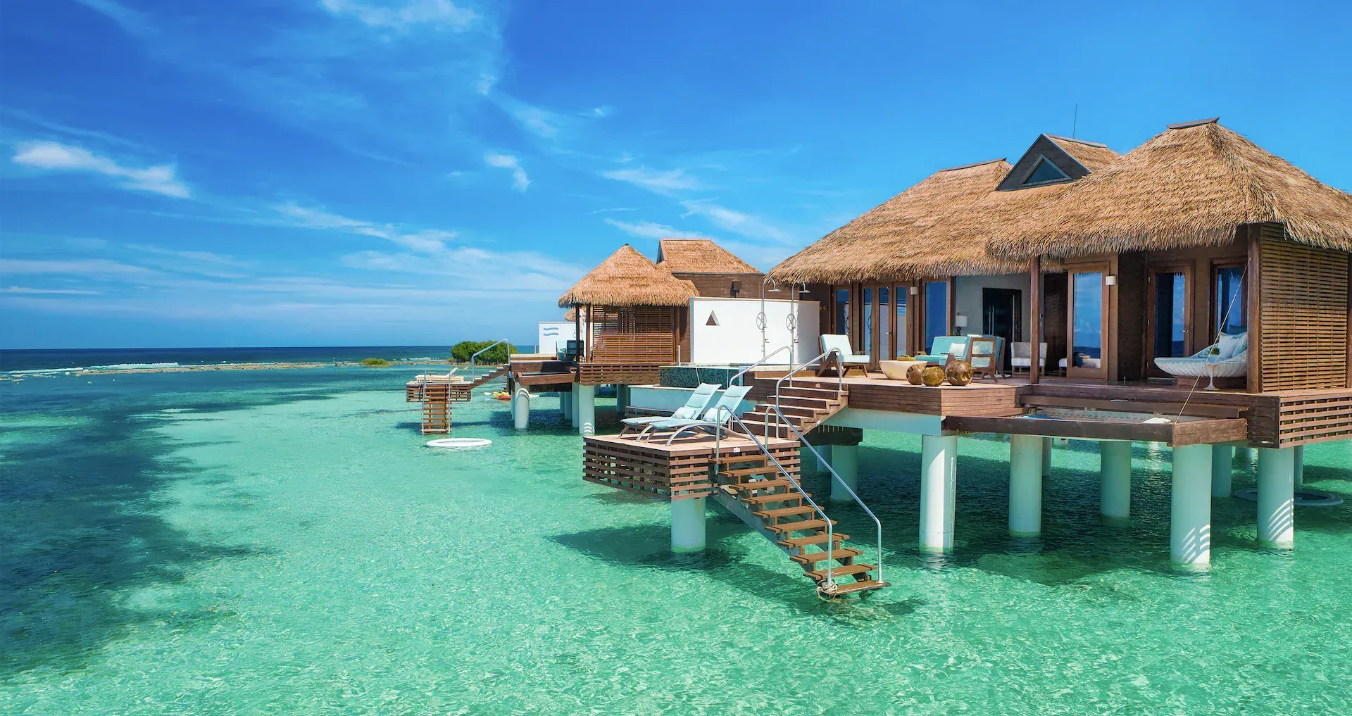 Overwater bungalows with thatched roofs standing on stilts in clear, turquoise tropical waters under a blue sky.