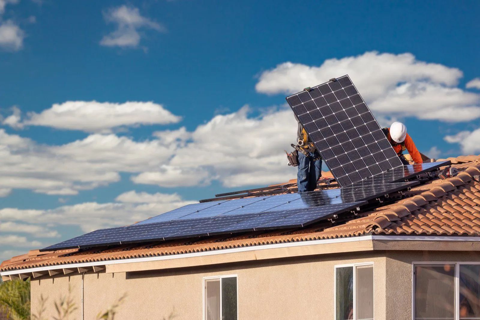 Two workers in safety gear installing solar panels on a house roof under a blue, cloudy sky.