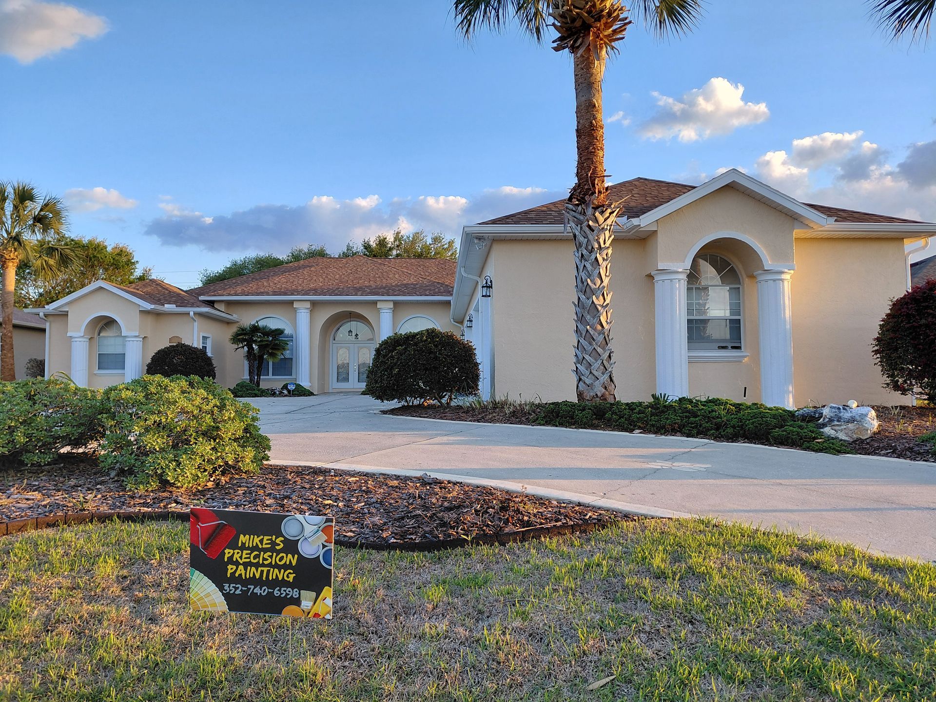 Single-story cream house with arched entry, palm tree, and a landscaped front yard under a blue sky.