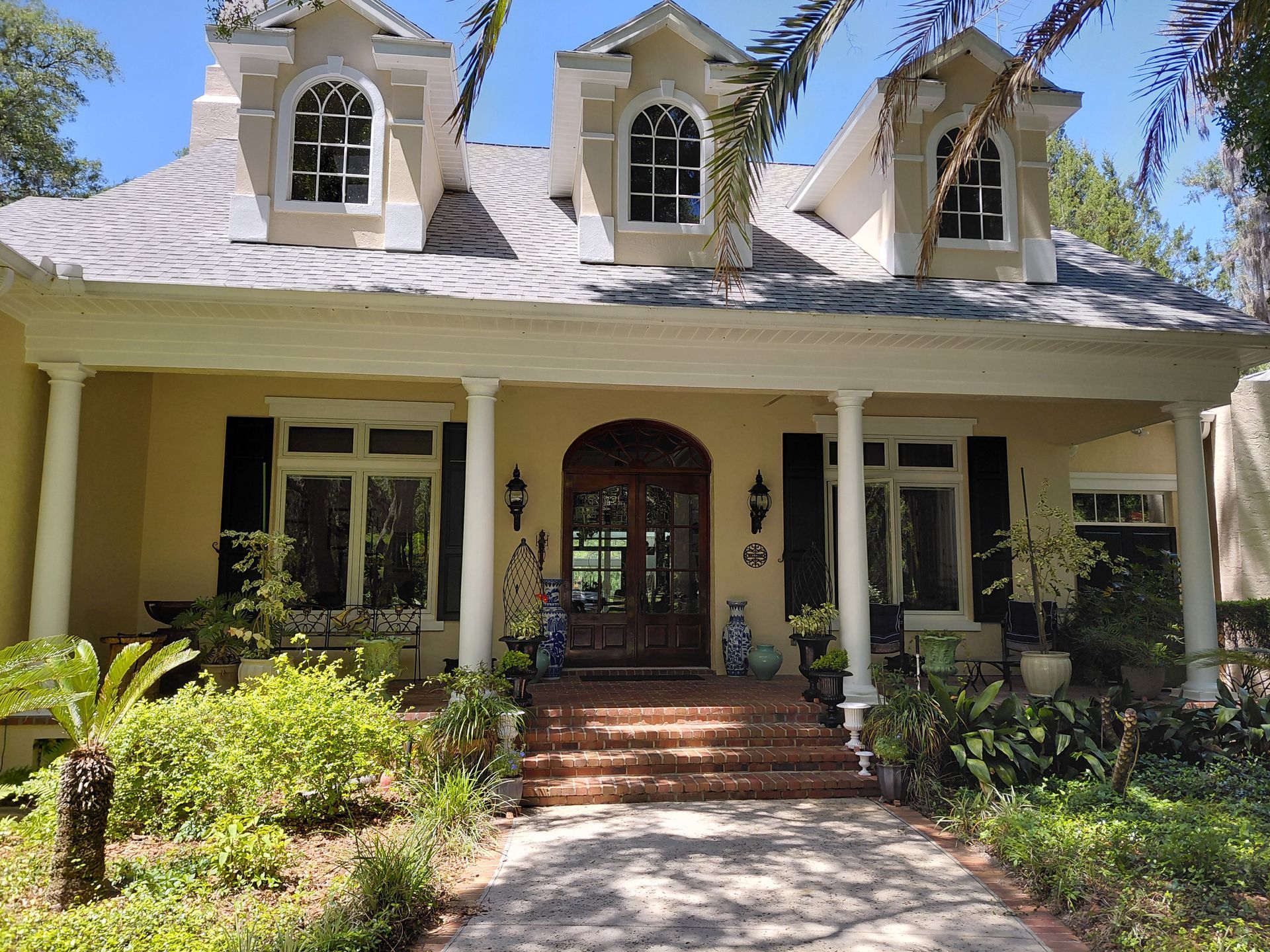 Historic cream-colored house with a front porch, brick steps, and dormer windows amid greenery