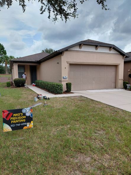 Single-story beige house with a garage, cloudy sky, and a yard sign in the front lawn.