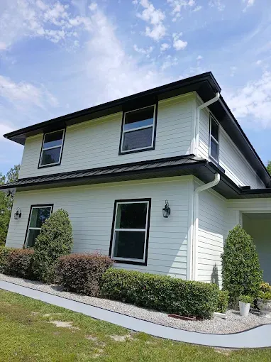 Two-story white house with black trim, windows, and roof. Bushes and green lawn in front. Blue sky.