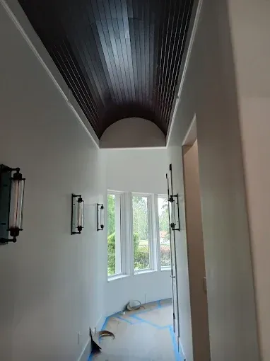 Hallway with a dark wood plank ceiling, white walls, arched opening, windows, and sconce lighting.