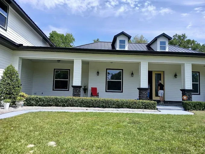 White house with black trim, dormers, and a covered porch; person stands in doorway.
