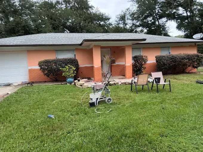 House with orange and white paint, lawn, power washer, bushes, and chairs on the grass.