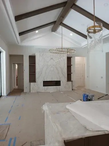 Interior view of a living room under construction, with marble fireplace, shelves, and wood beams on the ceiling.