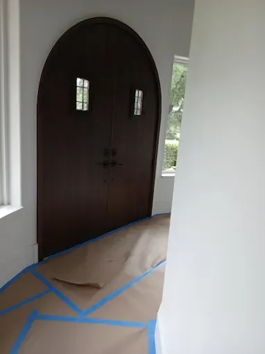 Arched brown double doors with small windows, hallway with floor covered for painting, white walls.
