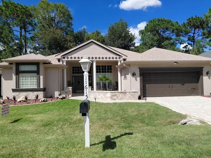 Beige ranch-style house with green lawn, mailbox, and trees against a blue sky with clouds.