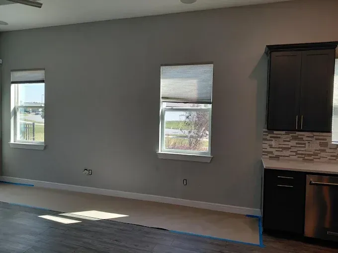 A gray-painted wall with two windows and dark cabinets in a room with blue painter's tape on the floor.