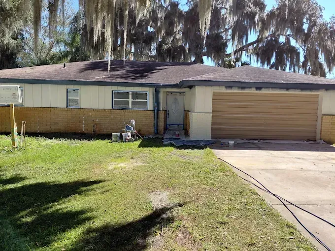 Ranch-style house with tan siding, brown garage door, and a green lawn. Trees hang in the background.