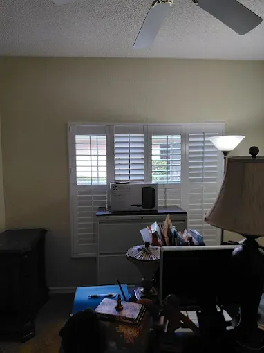 Office interior with window shutters, desk, lamp, and ceiling fan. Light-colored walls and furniture.