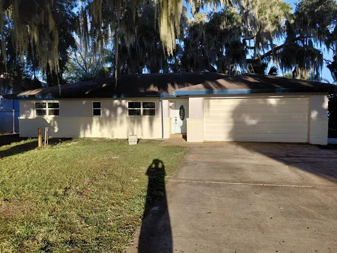 Ranch-style house with a light-colored exterior and a two-car garage. A long shadow falls on the driveway and lawn.