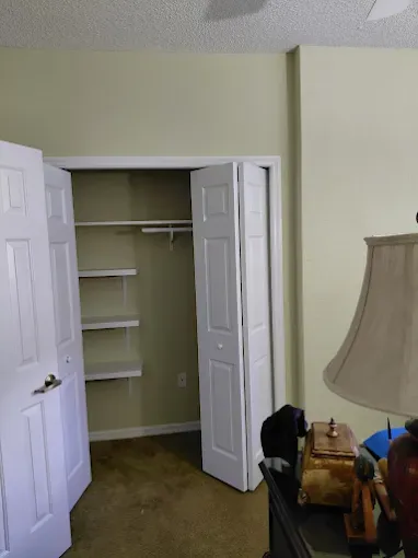 Closet with white bi-fold doors, shelves, and a rod. The walls are pale green, and there is a dark brown nightstand in the foreground.