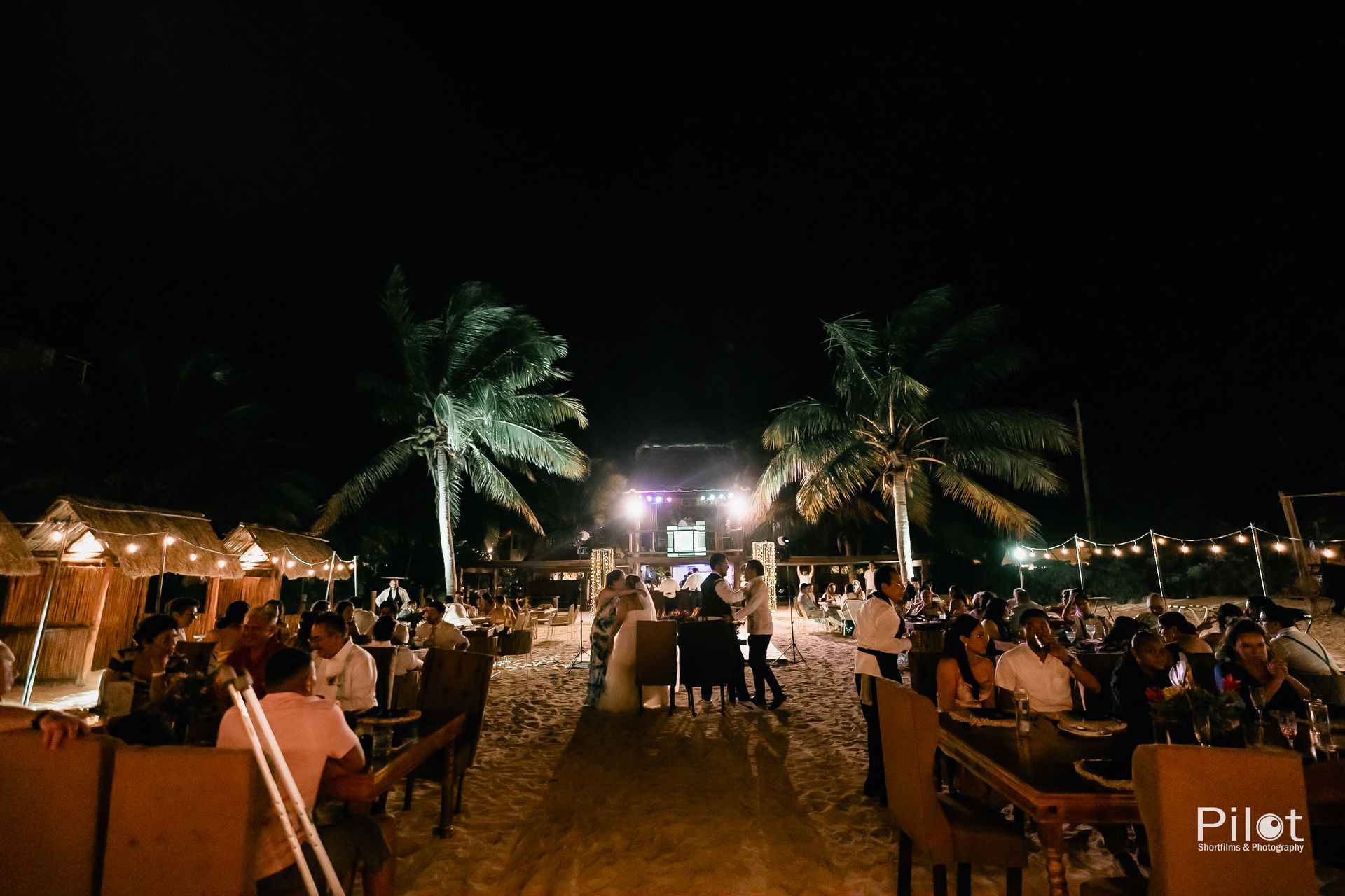 Un grupo de personas están sentadas en mesas en una playa por la noche.