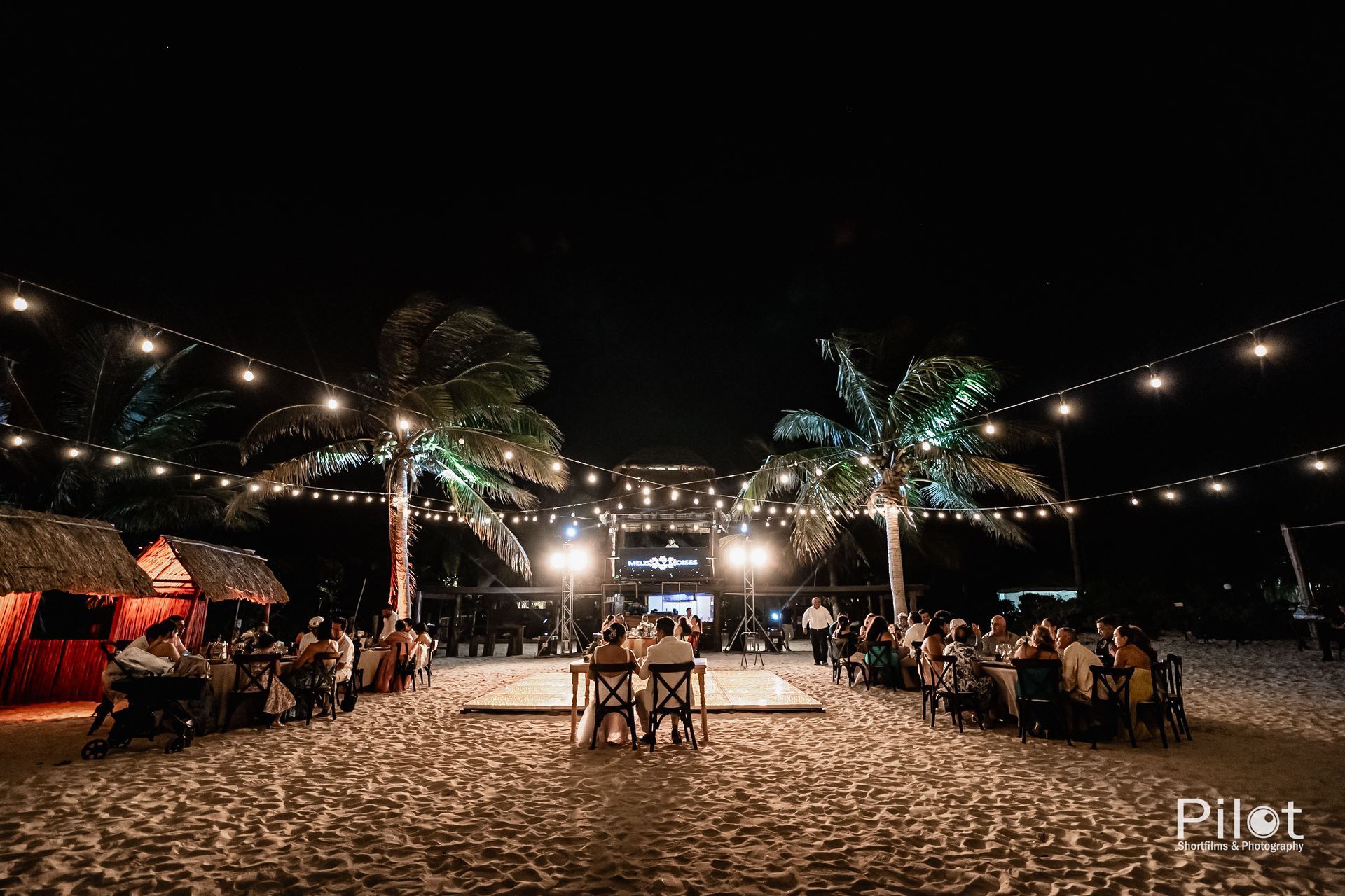 Un grupo de personas están sentadas en mesas en una playa por la noche.