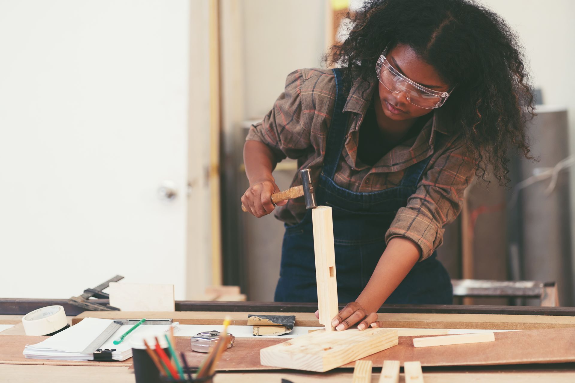 A person wearing protective goggles uses a hammer to assemble wooden pieces on a workbench in a workshop.