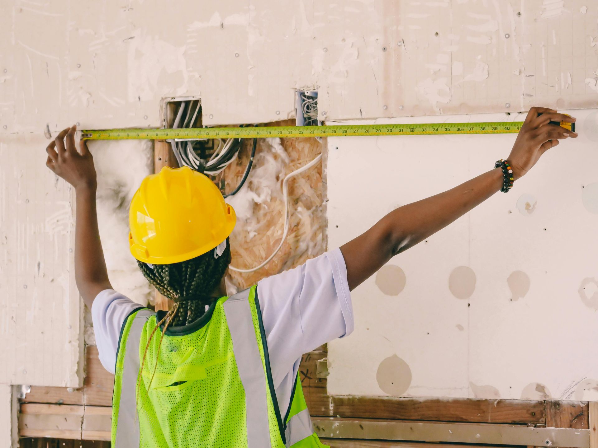 A construction worker in a yellow hard hat and neon vest uses a yellow tape measure to measure a wall opening.