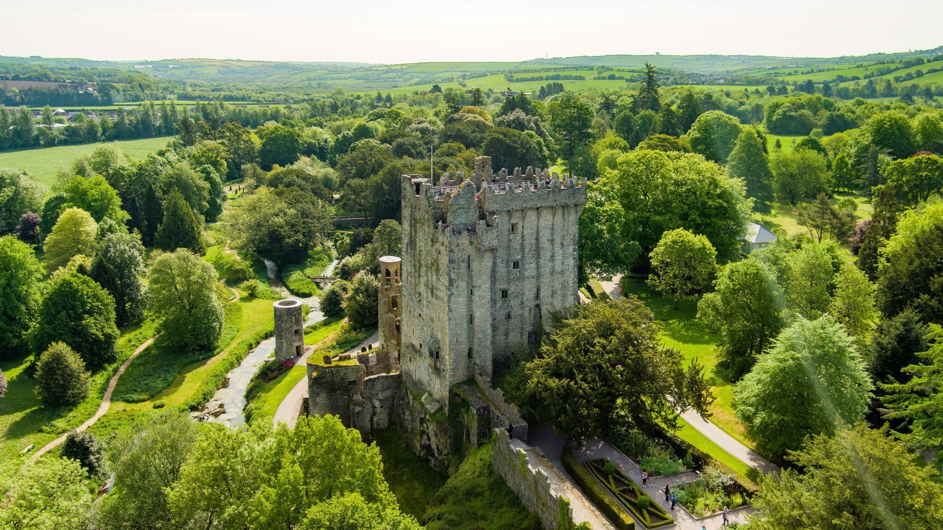Blarney Castle in Ireland, surrounded by lush green trees and gardens on a sunny day.