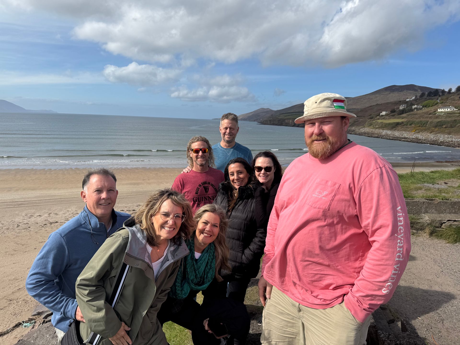 Group of eight people smiling at the camera on a beach, sunny day, ocean in background.
