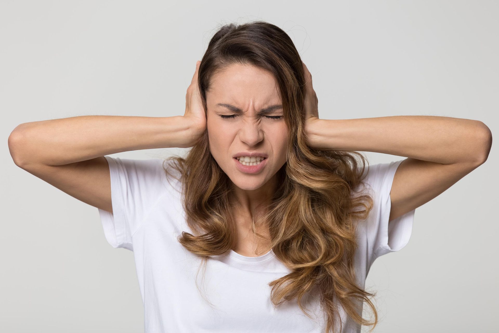 Woman with long hair covering her ears and grimacing in frustration against a plain background