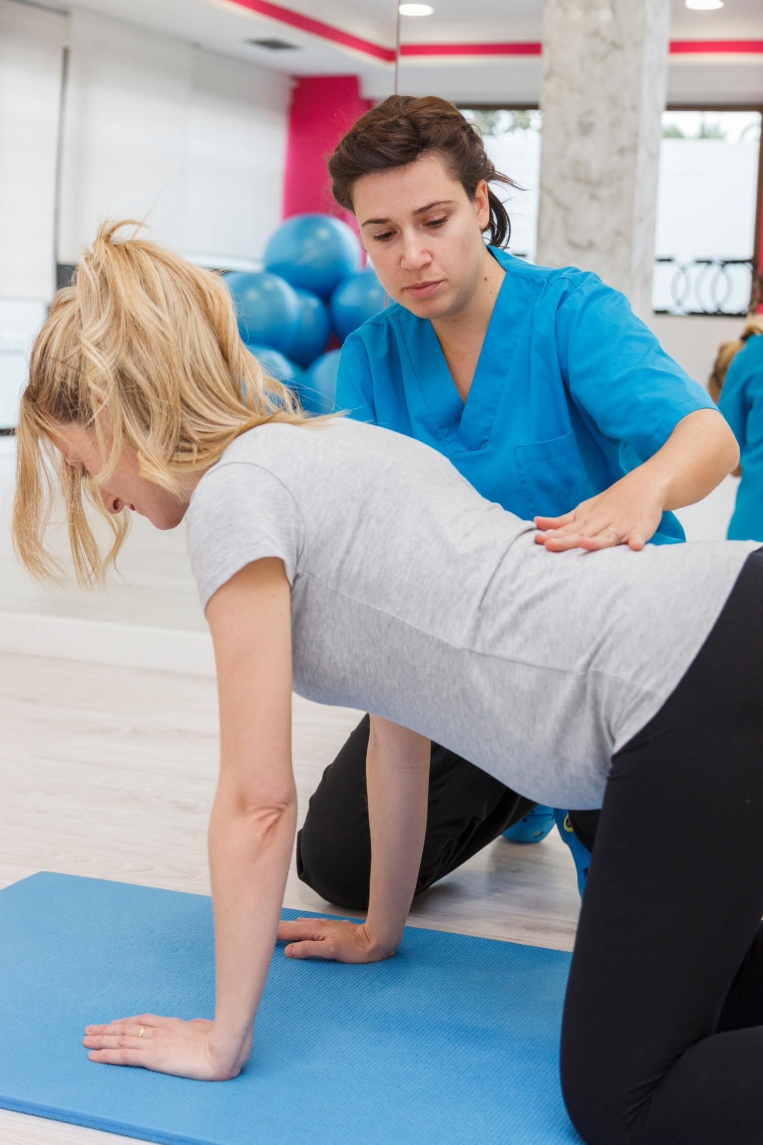 Physical therapist assisting a woman doing an exercise on a blue mat in a gym-like studio