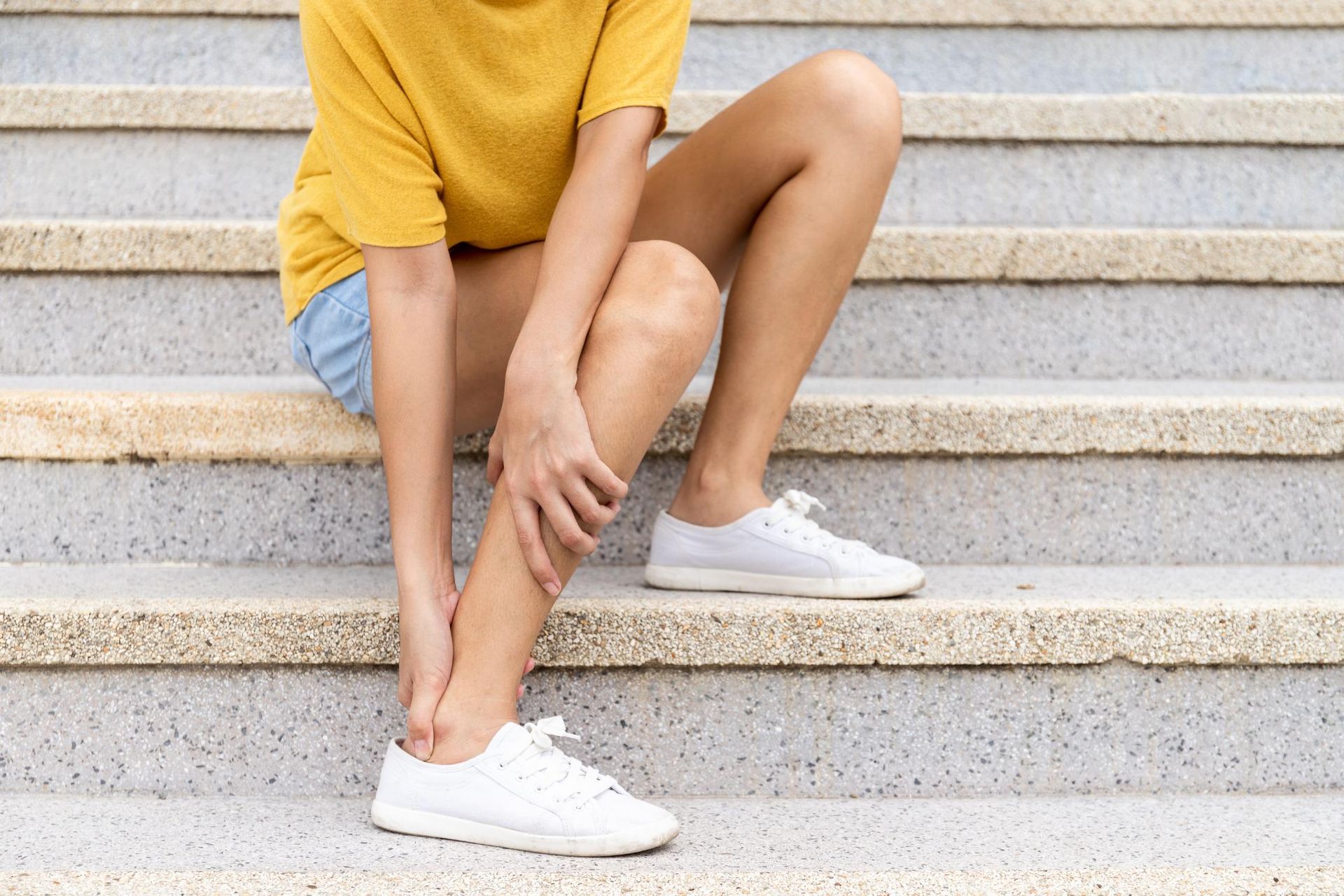 Person in a yellow shirt and denim shorts sitting on outdoor steps, holding one knee and wearing white sneakers
