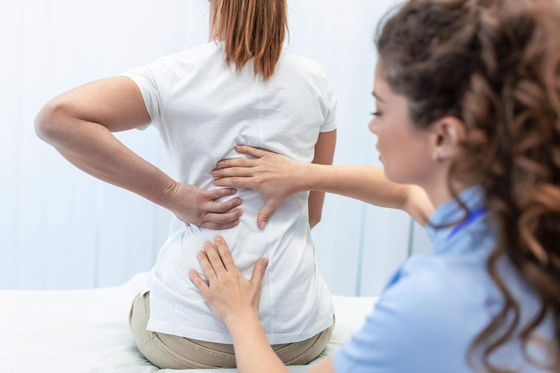 Physical therapist examining a patient’s back while she holds her lower spine in a bright clinic