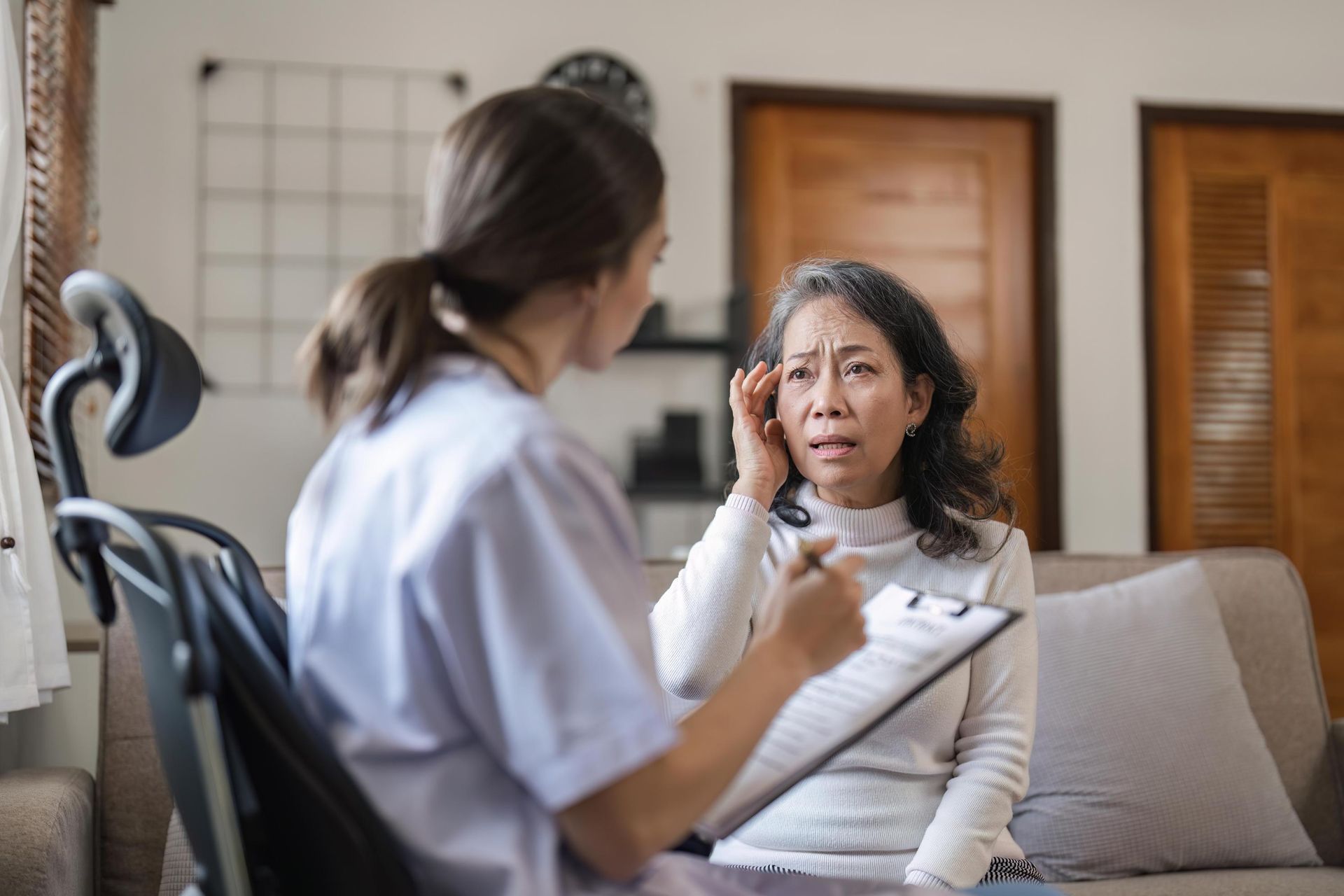 Therapist listening to a woman on a couch in a bright living room, holding a clipboard.
