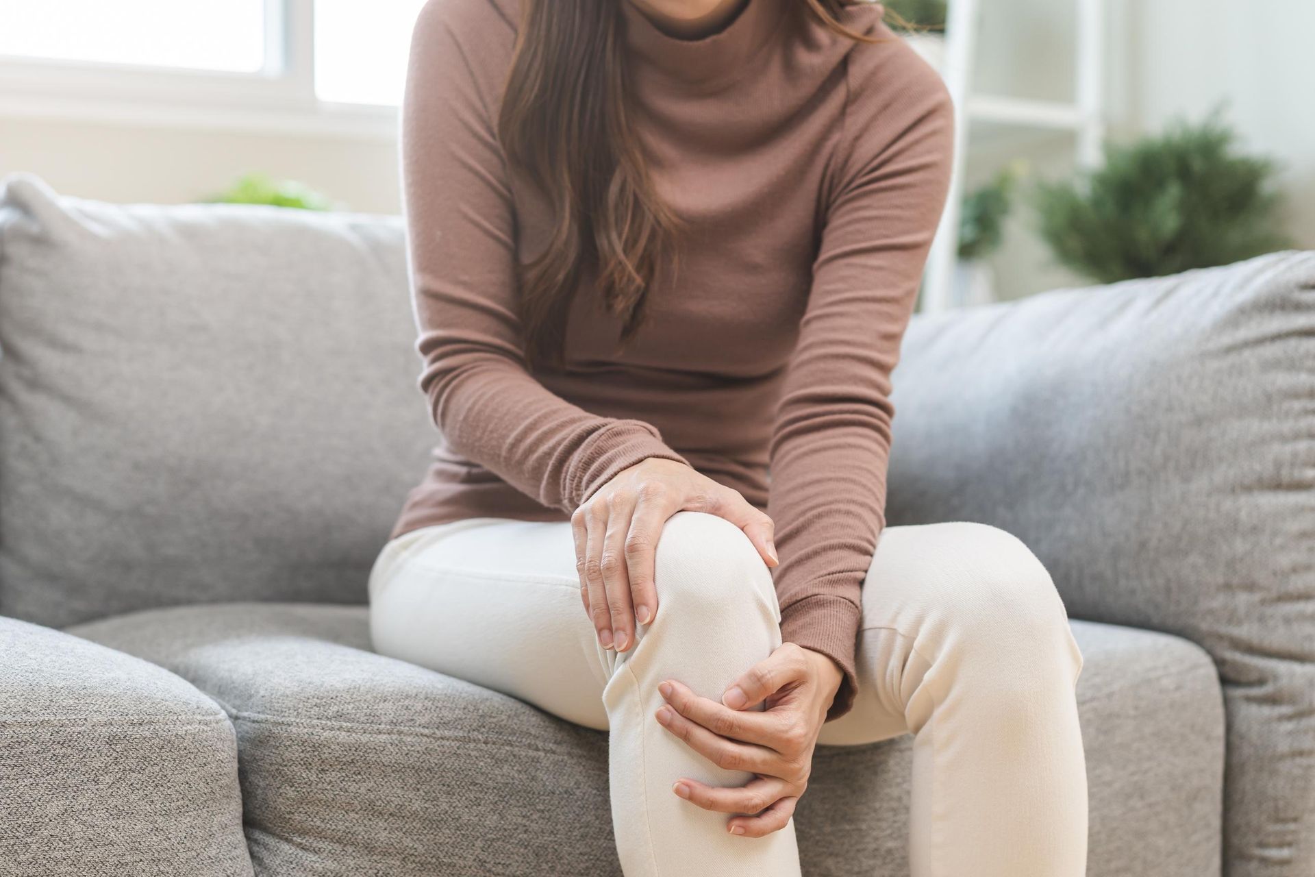Person sitting on a gray sofa, holding a knee with a hand, suggesting knee pain.