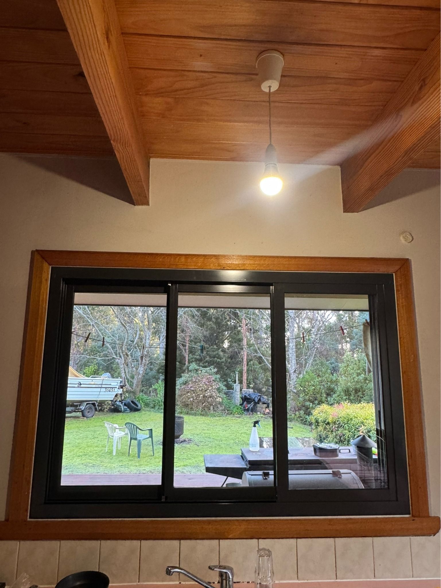 A dark-framed sliding window with wooden trim, set into a wall below a wooden ceiling beam and hanging light fixture. — Channel Glazing In Electrona, TAS