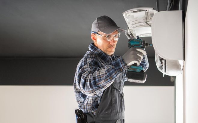 Electrician in plaid shirt and cap repairing a ceiling light fixture with tools.