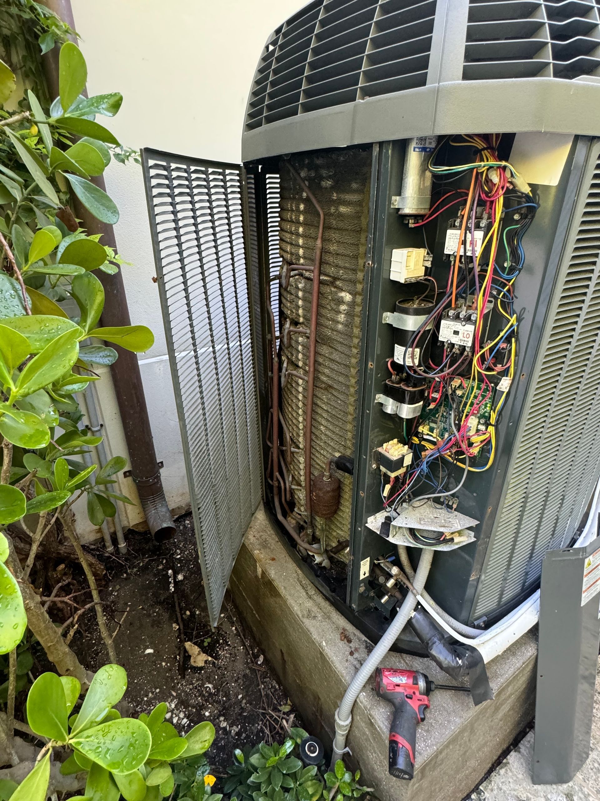 Outdoor air conditioner unit with open side panel, exposed wiring, and surrounding greenery