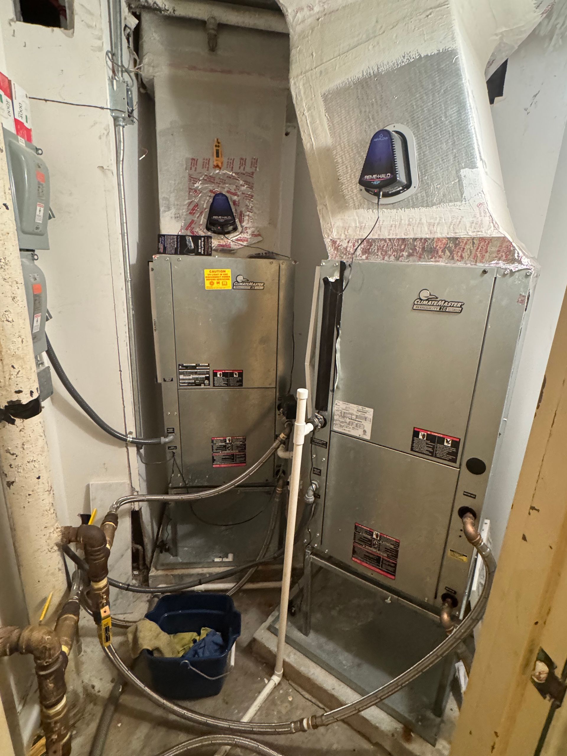 Electrical panels and pipes in a tight utility room with white insulation and a blue bucket on the floor