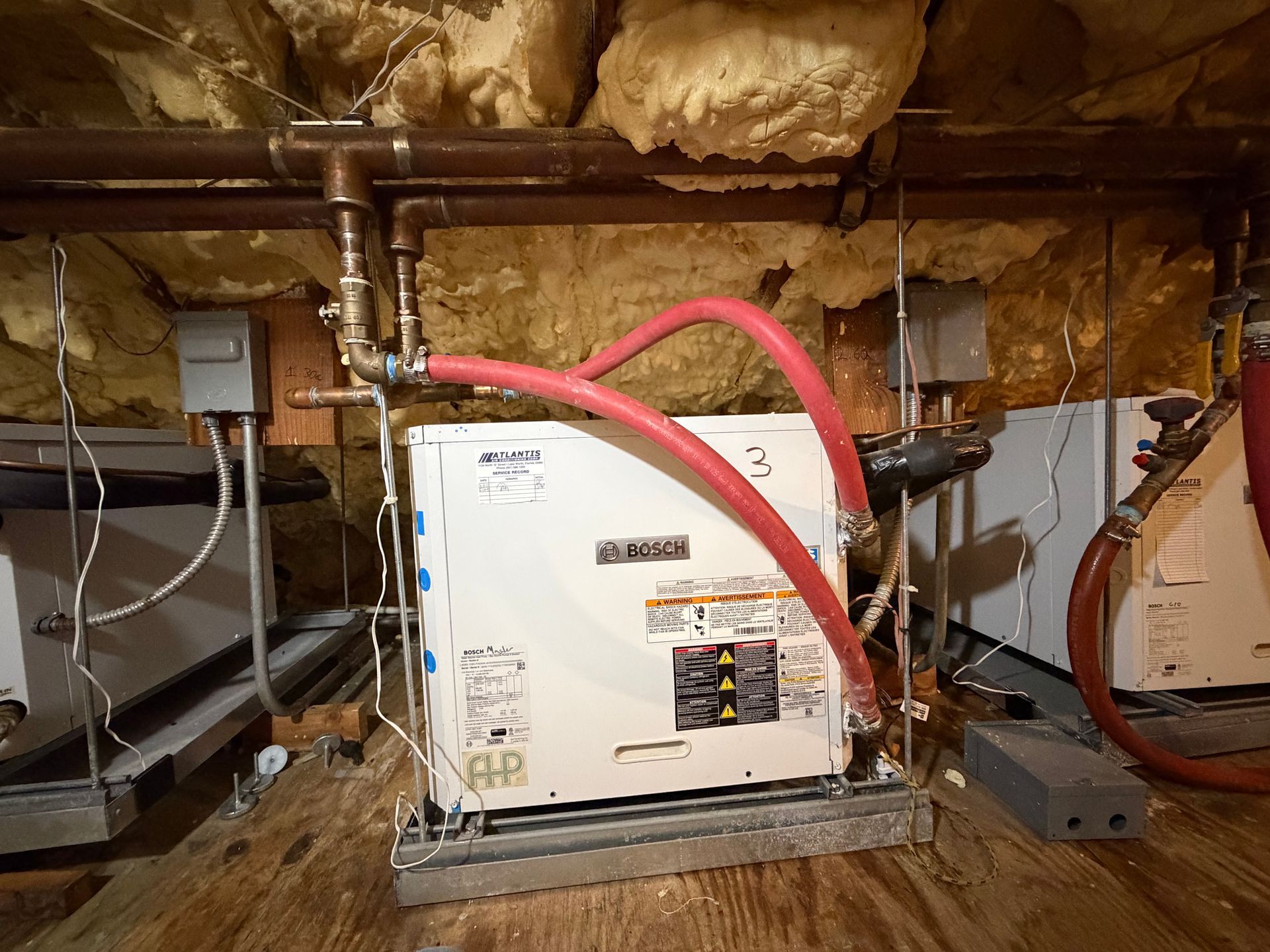 Central air handler in an unfinished attic with exposed insulation and ductwork, red hoses attached.