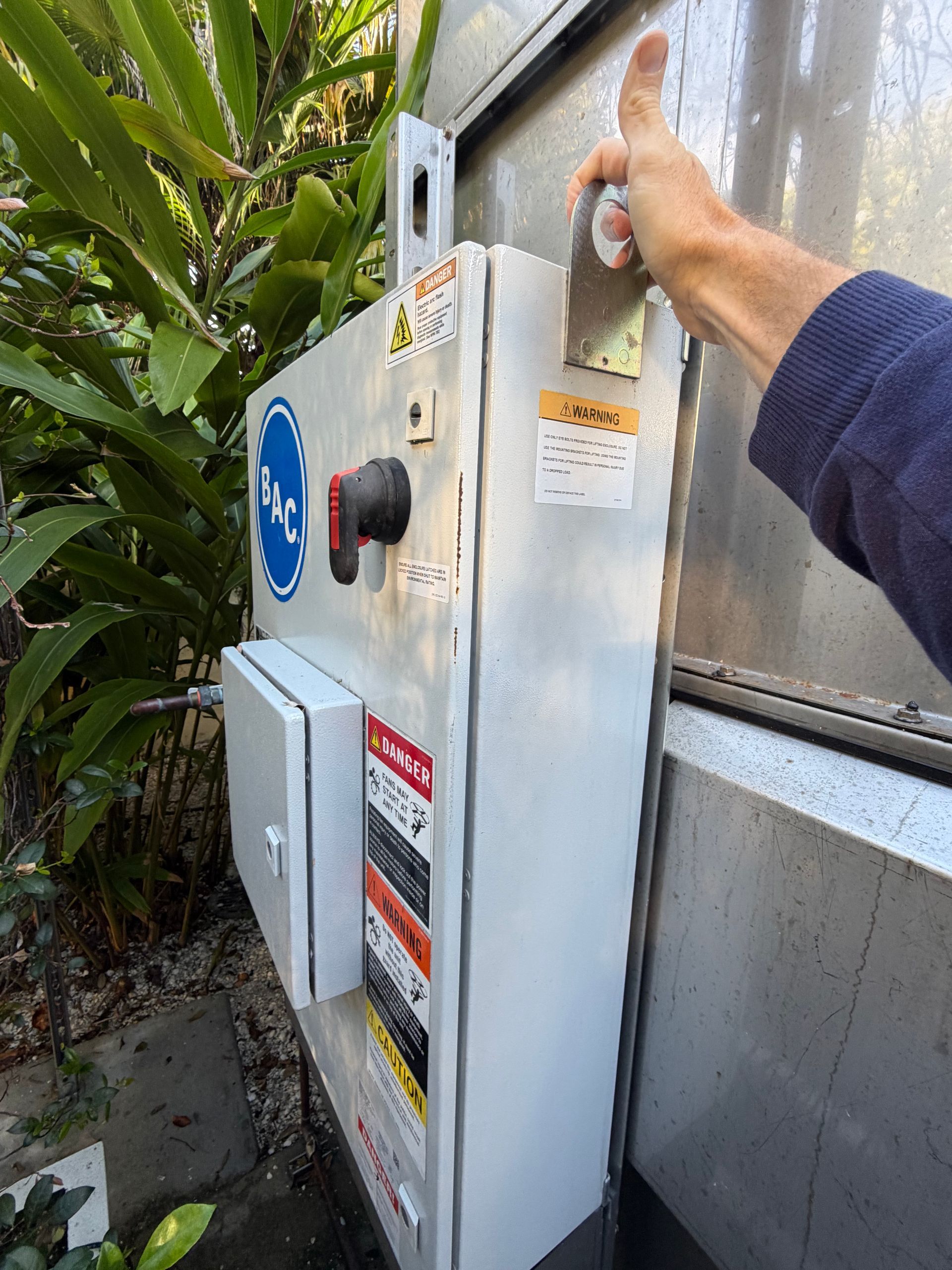 Person operating a large metal electrical control box outdoors beside greenery