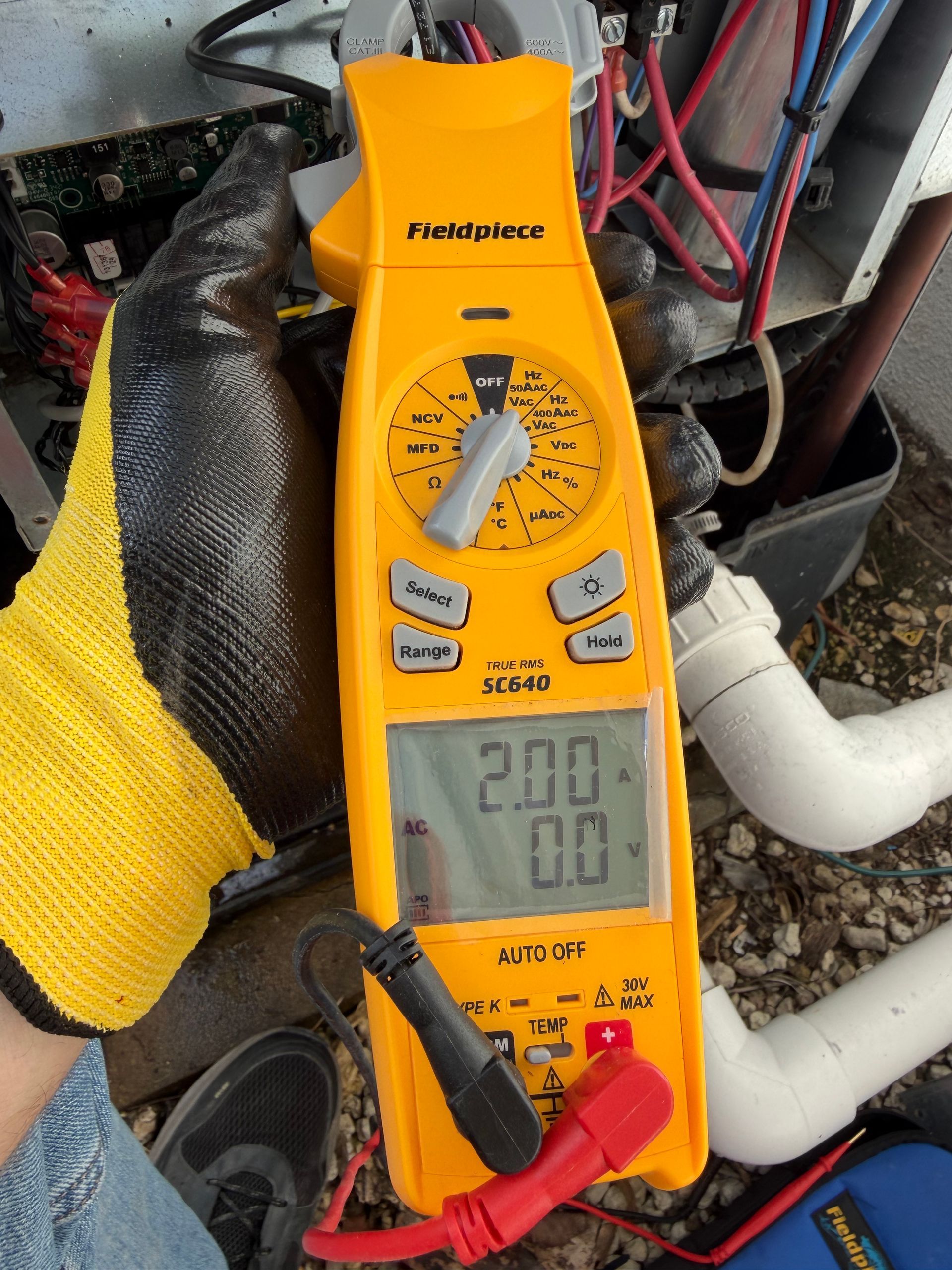 Yellow Fluke multimeter measuring a white pipe in a cluttered mechanical area.