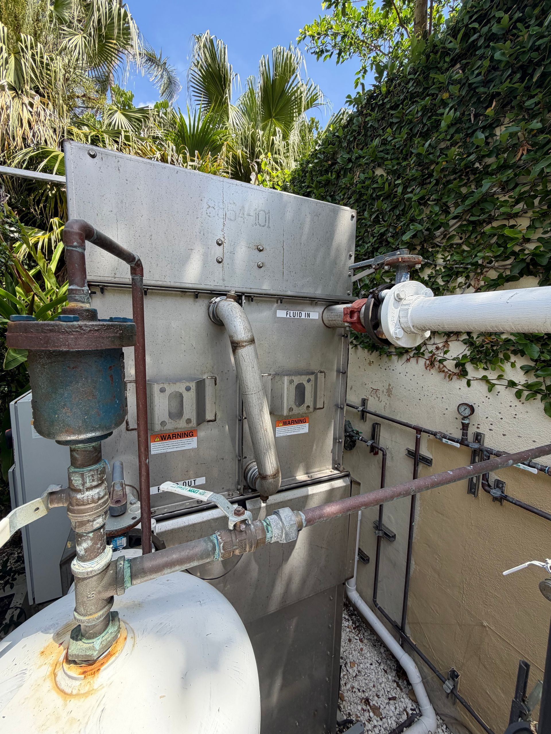 Outdoor industrial equipment with metal tanks, pipes, and valves beside a wall and greenery.