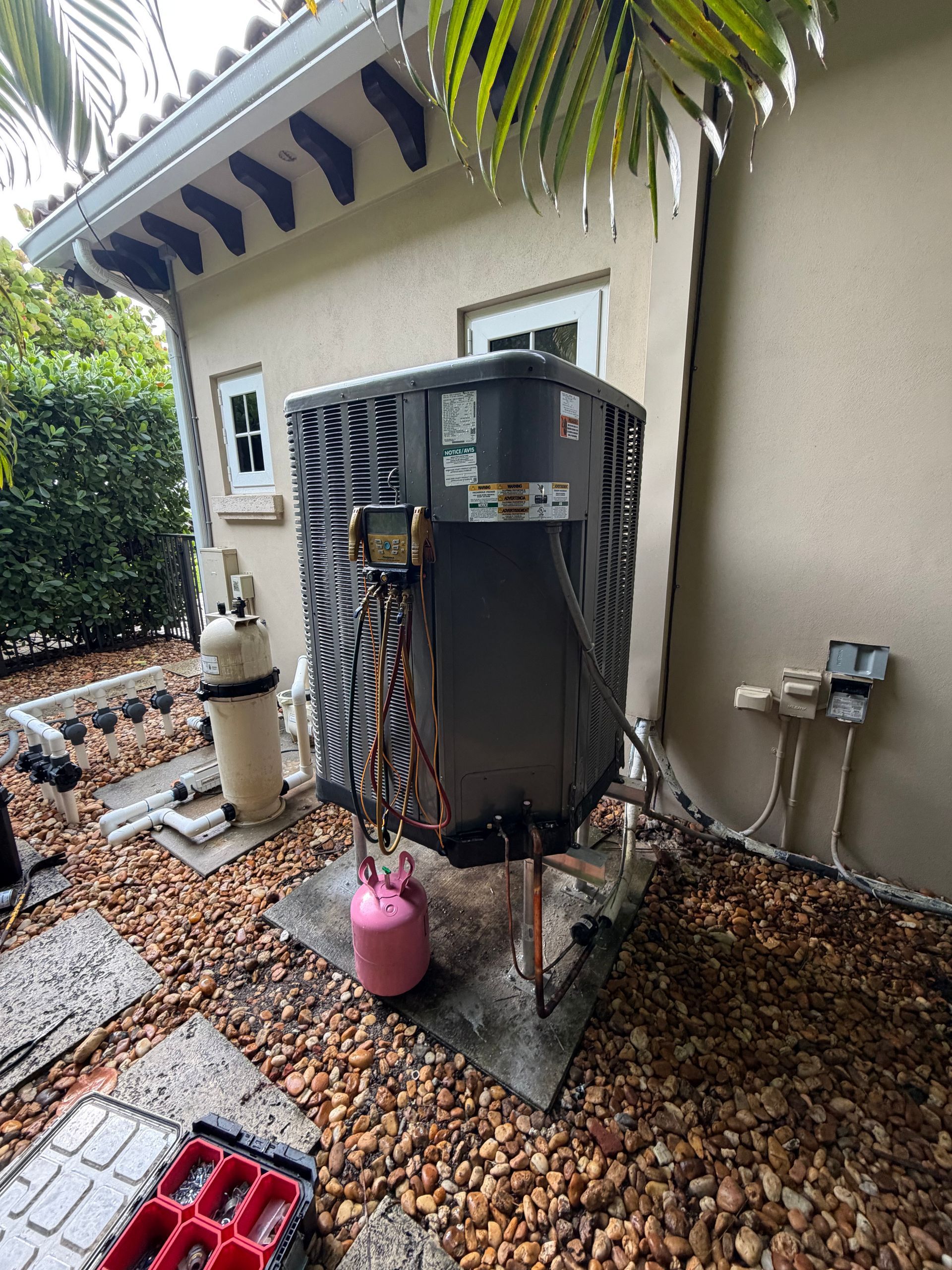 Outdoor HVAC unit with exposed pipes and a pink tank beside a house wall under a palm tree.