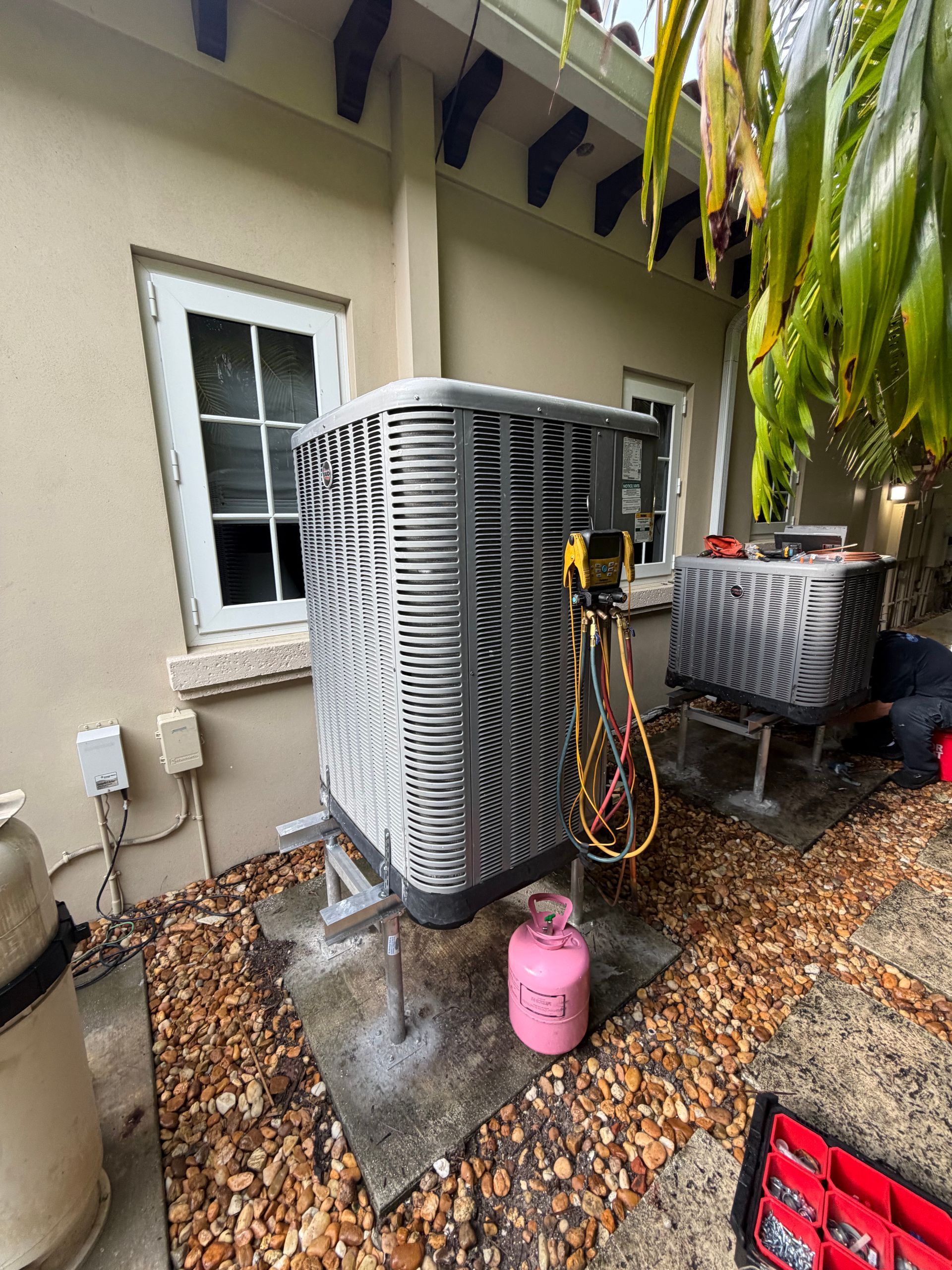 Two outdoor HVAC units beside a house, with hoses and a pink propane tank on a leaf-covered patio.